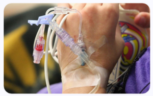 Mano de paciente con suero intravenoso insertado y sujeto con cinta adhesiva, mostrando tubulación médica colorida en un hospital.